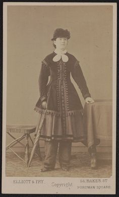 an old fashion photo of a woman in dress and hat with cane standing next to a table