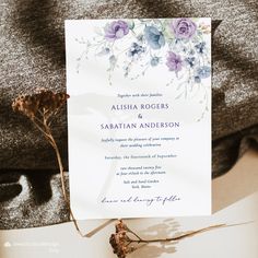 a white and blue floral wedding card on top of a table next to some dried flowers