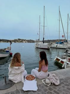 two women sitting on the dock eating pizza and watching boats in the water at dusk