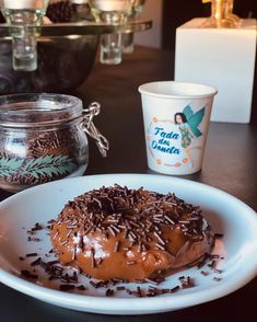 a chocolate donut with sprinkles sits on a plate next to two cups