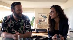 a man and woman sitting at a table with water in front of them, talking to each other