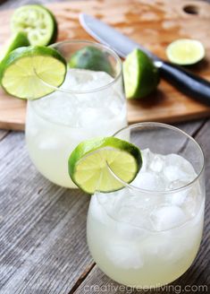 two glasses filled with ice and limes next to a cutting board