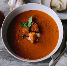 a bowl of tomato soup with bread on the side and garlic sprigs in the background