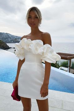 a woman standing next to a pool wearing a white dress with ruffles on it