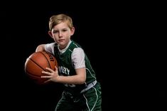 a young boy holding a basketball in his right hand and wearing a green uniform with white piping