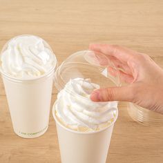 two cups with whipped cream in them on a wooden table, one being held by a hand