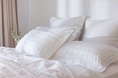 a white bed topped with lots of pillows and blankets next to a window covered in curtains