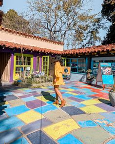 a woman in yellow shirt and shorts walking on tiled walkway next to colorful building with trees