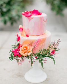 a three tiered cake with pink icing and orange flowers on the top is sitting on a white pedestal