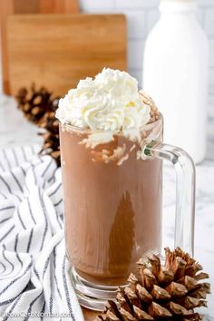 a glass mug filled with hot chocolate and whipped cream next to a pine cone on a table