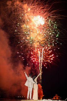 two people are standing on a dock with fireworks in the sky behind them and one person is raising their arms up