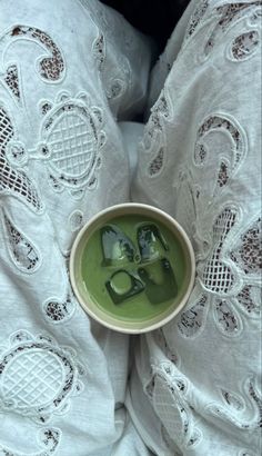 a bowl filled with green liquid sitting on top of a white bed cover covered in lace