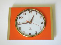 an orange and gold clock on a white wall with red trim around the hands, showing five o'clock