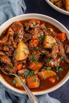 a bowl of beef stew with potatoes and carrots on a blue towel next to a spoon