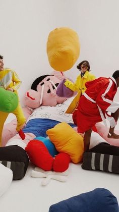 four men in bright colored clothing standing around a pile of pillows and stuffed animals on the floor