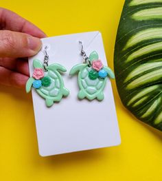 These cute Sea Turtle Earrings are handmade from polymer clay, resulting in a unique and eye-catching accessory. The earrings feature a beautifully painted shell design with delicate floral and succulent decorations in a mixture of pastel colours. Measuring approximately 3 cm in length, these earrings are the perfect size to make a statement. Despite their detailed design, they are remarkably lightweight, ensuring comfortable wear throughout the day. To add a touch of elegance and durability, ea Sea Turtle Earrings, Beachy Shell-shaped Earrings For Gift, Green Flower-shaped Earrings For Beach, Green Flower-shaped Beach Earrings, Summer Ocean-inspired Shell-shaped Earrings, Beach Lover Gifts, Alternative Jewelry, Succulents Decor, Beach Themed Party