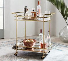 a gold bar cart with drinks on it in a living room next to a potted plant