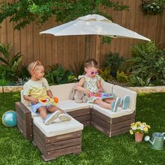 two children sitting on outdoor furniture with an umbrella over their heads and toys in the grass