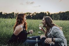 two women sitting on the grass drinking wine and talking to each other with trees in the background