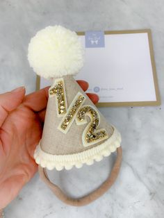 A handmade party hat perfect for marking 6 months of being born! Half birthday celebrations! A natural shade linen, with cream/off white wool pompom, pompom trim finished with gold glitter 1/2 (6 months). Other colours available at request (this listing is for the natural linen/gold glitter). Mini party hat measures 10cm high x 7cm at the base. Please note they are all made by hand so measurements may vary. The party hat is super soft and lightweight. It's made using premium fabrics including linen, wool and glitter fabric. The underside of the hat (the part of the hat that will sit on babies head) is lined with wool felt and the band to secure the hat on babies head is super soft and stretchy so it will be comfortable to wear. This party hat can be made using the other colours featured in