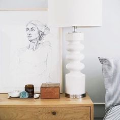 a nightstand with a lamp on it next to a painting and a bed in the background