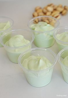 small cups filled with green cream sitting on top of a white table next to nuts