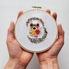 a person holding a small embroidered animal in front of a white wall with the image of a hedge on it