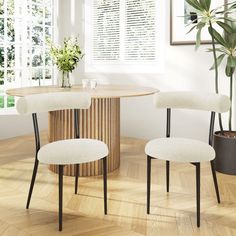 two white chairs and a round table in a room with wooden floors, plants and windows