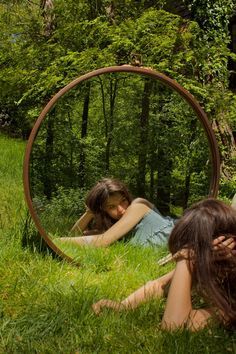 two women laying on the grass in front of a round mirror with trees behind them