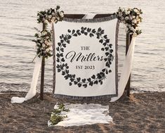 a wedding sign on the beach with flowers and greenery hanging from it's back