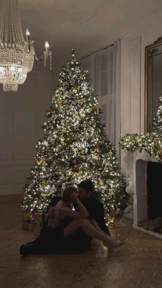 two people sitting on the floor in front of a christmas tree with lights and a chandelier