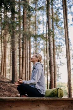 a woman sitting on top of a wooden bench in the woods doing yoga exercises with her eyes closed