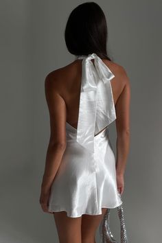 We recommend sizing down in this style. A major mini. The CARTIA is a soft Halter Micro Mini Dress that features an open back and statement bow detail to the neck band. It has been crafted from our lightweight satin fabrication, making it perfect for warm weather elopements or dance floor duties. Style with our Caitlin Diamante Rope Heels to keep it effortlessly elevated. FEATURES: High neckline Open back Large bow neck band detail A-line hem Lined Mini length FIT, FABRIC & CARE: Natalie is 173c Backless Dress Mini, Halter Bow Dress, Short Dress With Bow In Back, White Dress Purple Heels, Dress With Back Tie, White Vegas Dress, Mini Bow Dress, Short Wedding Reception Dresses, Satin Mini Dresses