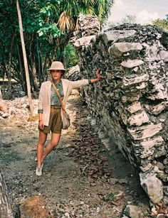 a woman walking down a dirt road next to a stone wall and palm trees in the background