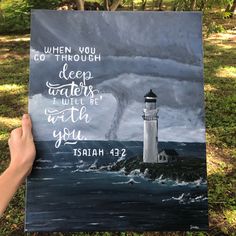 a hand holding up a painting with a lighthouse and bible verse on it in the grass