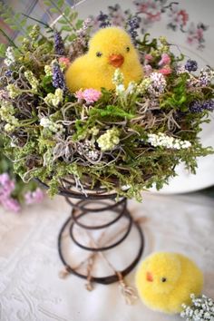 two small yellow chicks sitting in a basket filled with flowers and greenery on top of a table