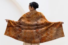 a woman is holding up an orange and brown shawl over her head while standing in front of a white wall