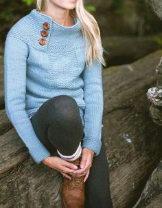 a woman sitting on top of a tree branch wearing a blue sweater and hat with buttons