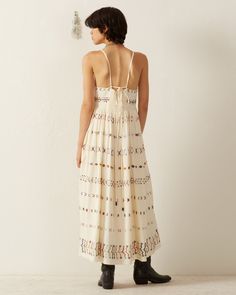 Crafted in Tencel, our ivory sleeveless dress is intricately hand-embroidered throughout with colorful stitching. The lightweight summer dress has a long gathered skirt and a beautiful drape. Lined in cotton, the dress has a cloth-covered button front, adjustable straps, and a smocked back for a comfortable fit. A classic silhouette perfect for warm weather. This one-of-a-kind textile was embroidered by hand. Subtle variations are part of the unique beauty of the garment and a signature of the a Sleeveless Dress With Smocked Back For Gatherings, Beige Cotton Dresses With Smocked Back, White Bohemian Dress With Smocked Bodice, Cream Embroidered Summer Beach Dress, Cream Cotton Sundress Maxi Dress, Cream Cotton Maxi Sundress, Summer Embroidered Cream Maxi Dress, Embroidered Cream Maxi Dress For Summer, Sleeveless Embroidered Beige Dress