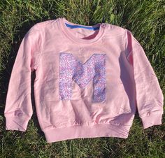 a pink sweatshirt with the letter m printed on it laying in the grass next to a pair of scissors