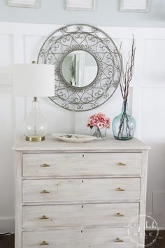 a white dresser topped with a mirror and vase filled with flowers next to a lamp