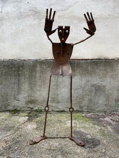 a metal sculpture with hands and legs on it's body, standing in front of a concrete wall