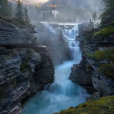 a waterfall in the middle of a forest with fog hanging off it's sides