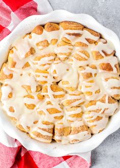 a white dish filled with cinnamon rolls covered in icing on top of a red and white checkered table cloth