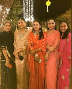 four women dressed in sari posing for a photo with lights hanging from the ceiling behind them