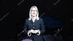 a woman with glasses standing in front of a black background wearing a suit and holding her hands together