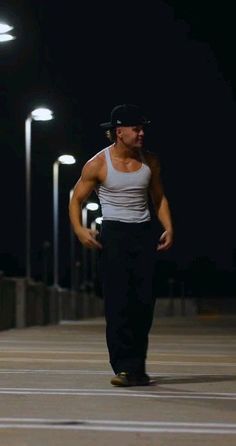 a man in black pants and white tank top walking down the street at night with his hands on his hips