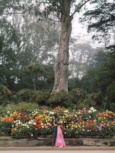 two people standing in front of a large tree and flower garden with flowers all around them