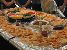 a table full of chips and dips with people in the background looking at them