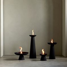 three black candle holders with flames in them on the floor next to a white wall